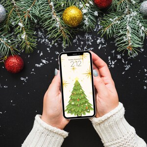 Puede incluir: Un tel&eacute;fono inteligente que muestra un fondo de pantalla de &aacute;rbol de Navidad es sostenido por una persona que usa un su&eacute;ter blanco. La pantalla del tel&eacute;fono muestra la hora 2:12 y la fecha domingo 10 de noviembre. El fondo incluye adornos navide&ntilde;os y ramas de pino.