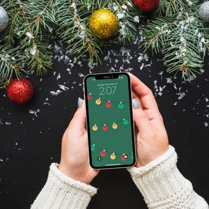 Puede incluir: Un tel&eacute;fono inteligente que muestra un fondo de pantalla de luces navide&ntilde;as, sostenido por una persona con un su&eacute;ter blanco. La pantalla del tel&eacute;fono muestra un fondo verde con luces de colores. La hora es 2:07. Adornos navide&ntilde;os y ramas de pino en el fondo.
