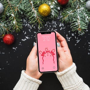 Puede incluir: Un tel&eacute;fono inteligente que muestra una pantalla rosa con un dise&ntilde;o de bast&oacute;n de caramelo, sostenido por una persona con un su&eacute;ter blanco. El fondo presenta adornos navide&ntilde;os y ramas de pino.