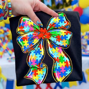 May include: Black t-shirt featuring a colorful puzzle piece bow design. The bow is outlined in gold glitter and filled with various colored puzzle pieces. The design is centered on the shirt, with a focus on autism awareness.