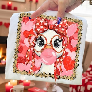 May include: White sweatshirt featuring a cartoon penguin with heart-shaped glasses and a bubblegum bubble. The penguin has a red bow with white hearts. The background is filled with pink and red hearts, and the border is gold.