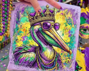 May include: A light purple t-shirt featuring a colorful illustration of a pelican wearing a gold crown and sunglasses, and a beaded necklace. The pelican is depicted with purple, green, and yellow feathers. The background has paint splatters in yellow, green, and purple.