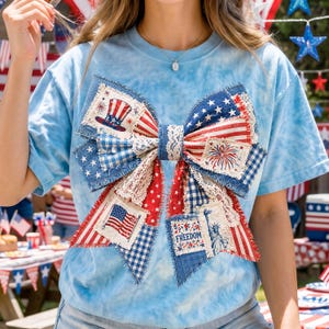 May include: A light blue tie-dye t-shirt featuring a large bow design. The bow is made of patches with patriotic themes, including the American flag, fireworks, and the Statue of Liberty. The word "FREEDOM" is also visible.