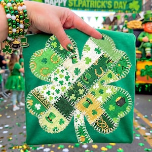 May include: Green t-shirt featuring a four-leaf clover design made of various green and white patterned fabrics. The clover includes shamrocks, leprechaun hats, and horseshoes. The shirt is held up in front of a parade with the text "HAPPY ST. PATRICK'S DAY" in the background.