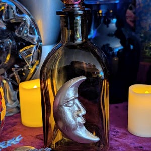 May include: A translucent amber glass bottle with a spherical stopper, featuring a prominent silver-toned crescent moon emblem with a human face relief attached to the front surface. The bottle has a rectangular base and a narrow neck design.