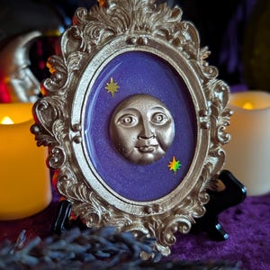 May include: An oval-shaped decorative frame in a metallic gold finish with ornate scrollwork detailing. The center features a smooth, deep purple resin inlay with a raised, three-dimensional gold moon face and two small, multi-colored star accents.