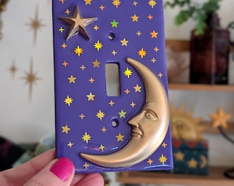 May include: A purple light switch cover with a gold crescent moon and star design. The cover is decorated with gold stars and a large gold star. The switch plate is held by a person with pink nail polish.
