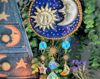 Könnte beinhalten: Ein Wandbehang mit Sonne und Mond im Himmel, mit blauem Hintergrund und goldenem Glitzer. Sonne und Mond sind silberfarben und haben Gesichter. Der Behang hat Kristall- und sternförmige Anhänger.