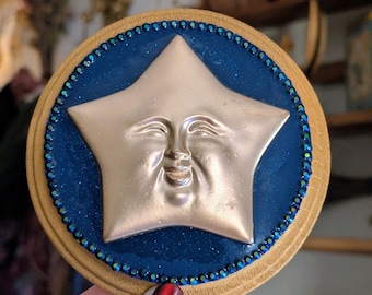 Könnte beinhalten: Runde Holzdekoration mit einem lächelnden Stern-Design. Der goldene Stern ist vor einem dunkelblauen, glitzernden Hintergrund platziert. Der Rand ist helles Holz mit einer Perlenverzierung.