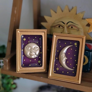 May include: Two framed celestial art pieces. One features a full moon face, the other a crescent moon face, both in gold against a purple starry background. The frames are gold. A wooden sun sculpture is in the background.