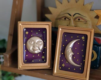 May include: Two framed celestial art pieces. One features a full moon face, the other a crescent moon face, both in gold against a purple starry background. The frames are gold. A wooden sun sculpture is in the background.
