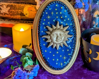 Könnte beinhalten: Ovale Wanddekoration mit goldenem Rahmen. Es zeigt einen blauen Hintergrund mit goldenen Sternen und einer goldenen Sonne mit Gesicht. Der Rahmen ist mit kleinen schwarzen Perlen verziert. Der Hintergrund ist tiefblau.