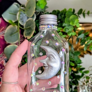 Puede incluir: Una botella de vidrio transparente con tapa plateada y un diseño de luna sonriente. La botella está decorada con pedrería iridiscente y una luna plateada. La botella se sostiene en una mano.