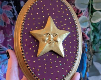 Könnte beinhalten: Ovale Wanddekoration mit goldenem Rahmen. Die Mitte hat einen tiefvioletten Hintergrund mit goldenen Sternen und einem goldenen Stern mit einem lächelnden Gesicht. Der Rand ist mit kleinen, irisierenden Perlen verziert.