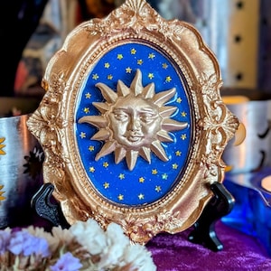 May include: An ornate, gold-coloured framed wall art piece. The oval centre has a blue background with gold stars and a gold sun with a face. The frame is also gold and has decorative detailing. The art piece is displayed on a black stand.