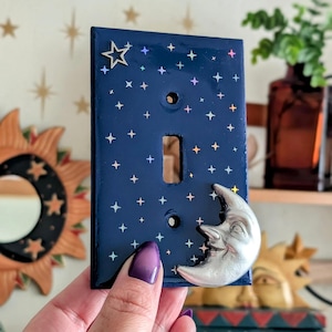 May include: A navy blue light switch cover decorated with a celestial theme. It features a silver crescent moon and star accents. The cover is adorned with iridescent star details, adding a touch of sparkle.