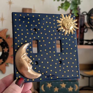 May include: A navy blue double light switch cover decorated with gold stars. The cover features a gold sun and crescent moon design. The switch plate is a decorative home decor item.