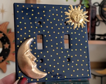 May include: A navy blue double light switch cover decorated with gold stars. The cover features a gold sun and crescent moon design. The switch plate is a decorative home decor item.