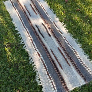 Puede incluir: Una alfombra rectangular tejida a mano con un patrón de rayas grises y marrones. La alfombra presenta rayas alternas de gris claro, gris oscuro y marrón, con elementos decorativos blancos y marrones. Los bordes tienen un acabado con flecos.