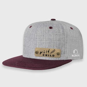 May include: A gray and burgundy baseball cap with a gold metal plate that says "wild child" with a feather design. The cap has a small logo on the back that says "RUBDE".