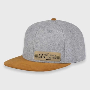 May include: A gray and brown baseball cap with a metal plate that reads "BESTER PAPA ALLER ZEITEN".