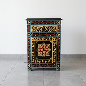 May include: A rectangular wooden bedside table featuring a black base with intricate hand-painted patterns in yellow, red, and light blue. The cabinet includes one top drawer and a single door with a central star-shaped geometric motif.