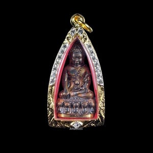 May include: A gold pendant with a red and gold frame, featuring a small, dark brown Buddha statue. The frame is decorated with clear rhinestones.