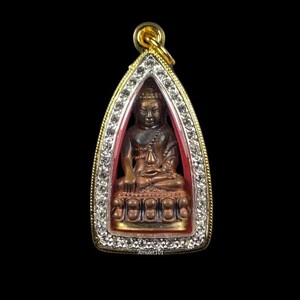 May include: A gold pendant with a small, brown Buddha statue inside. The pendant is surrounded by a clear, red, and white frame with small, clear stones around the edge.