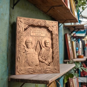 Pode incluir: Um ícone de madeira esculpida que representa dois santos, com detalhes intrincados e um fundo dourado. O ícone é emoldurado em uma moldura simples de madeira e apresenta o texto "Святой великомученик и страстотерпец и Игнат" em escrita cirílica.