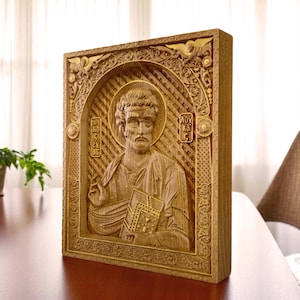 Op de afbeelding: Een rechthoekige houten plaquette met een gedetailleerd reliëf van een bebaarde figuur die een boek vasthoudt. De centrale figuur is omlijst door een boogvormige rand met ingewikkeld krulwerk en tekst in Griekse tekens aan weerszijden van de halo.