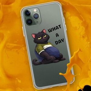 May include: A clear smartphone case featuring a cartoon illustration of a grumpy black cat wearing a green and blue shirt, with the text WHAT A DAY printed in black. The case is surrounded by a vibrant, thick splash of bright orange liquid.