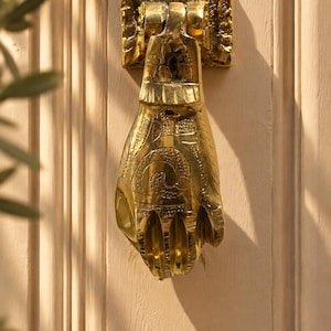 May include: A polished brass door knocker shaped like a human hand, featuring intricate engraved patterns on the wrist and palm area. The hand is mounted on a rectangular textured base plate and hangs vertically against a light-colored door.