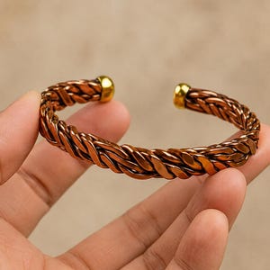 Op de afbeelding: Een koperkleurige manchetarmband met een gevlochten metalen ontwerp. De armband heeft een gepolijste, reflecterende afwerking en is aan beide open uiteinden voorzien van gladde, afgeronde, goudkleurige metalen uiteinden die contrasteren.