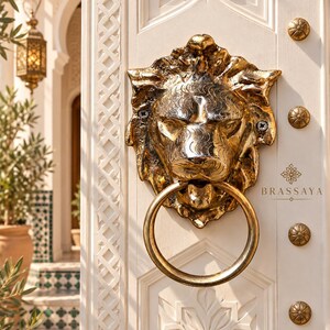 Könnte beinhalten: Ein goldfarbener Türklopfer in Löwenkopfform mit einem kreisförmigen Ring, montiert an einer weißen Holztür mit aufwendigen geschnitzten geometrischen Mustern und kleinen runden goldenen Nieten. Der Text BRASSAYA steht auf der Tür.