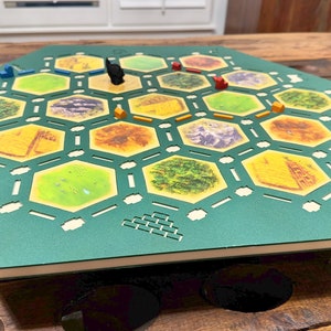 May include: A green hexagonal board game with various terrain tiles, including forests, mountains, and fields. The game board has small wooden pieces in red, blue, black, and orange. The board is set on a wooden table.