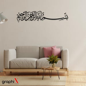 Op de afbeelding: Zwarte Arabische kalligrafie muurkunst met elegant schrift. Het kunstwerk is gemonteerd boven een lichtgrijze bank met roze kussens, een houten salontafel en een vaas met groen. De kamer heeft een neutraal kleurenpalet.