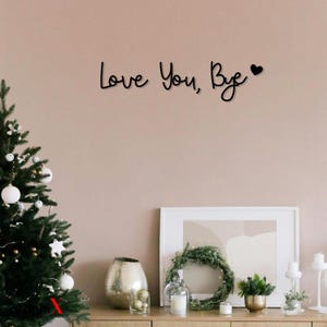 May include: A black metal wall decor with the text "Love You, Bye" and a small heart symbol. The decor is hanging on a light pink wall above a wooden table with a Christmas tree, a wreath, and decorative items.