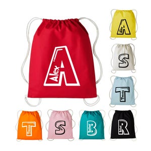 May include: A selection of drawstring bags in different colours, each with a large letter and a name. The main red bag shows a white "A" with "Alex" inside. Other bags are yellow, cream, light blue, orange, pink, and black.