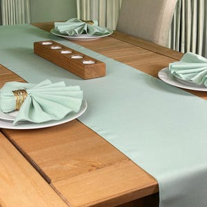 May include: A light green table runner with a wooden candle holder and folded napkins on a wooden table.