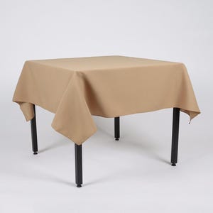 May include: A square, tan tablecloth draped over a table with black legs. The tablecloth has a smooth, even texture.