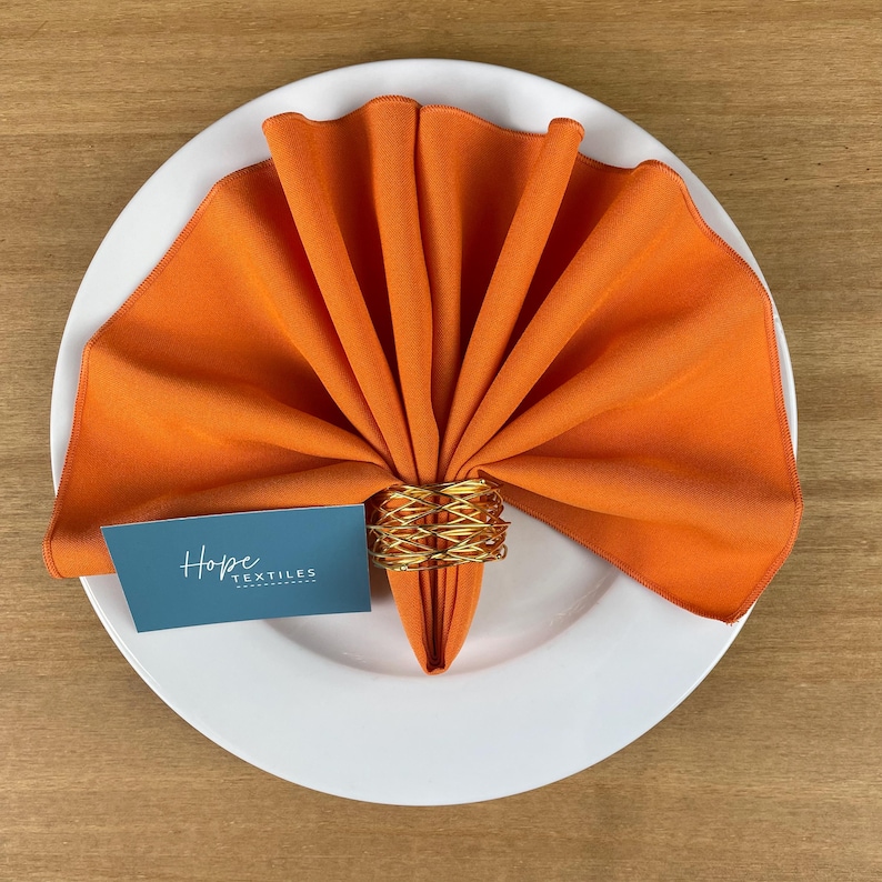 Pode incluir: Guardanapo de linho laranja dobrado em forma de leque e mantido junto com um anel de guardanapo de arame dourado. Um cart&atilde;o de visita com o texto "Hope Textiles" est&aacute; colocado no prato.