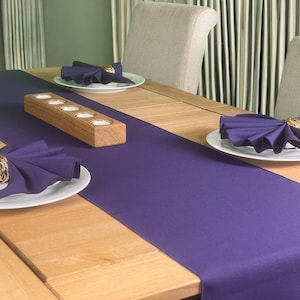 May include: A purple table runner with a wooden candle holder on a wooden table with white plates and purple napkins.