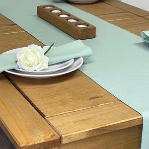 May include: A wooden table setting with a pale green table runner, napkins, and white plates. A wooden candle holder with four openings is on the runner. White roses are placed on the plates. The table is made of light brown wood.
