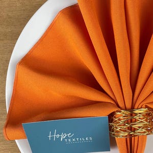 Pode incluir: Guardanapo de linho laranja dobrado em forma de leque e preso com um anel de guardanapo de arame dourado. O guardanapo est&aacute; sobre um prato branco com um cart&atilde;o que diz "Hope Textiles".