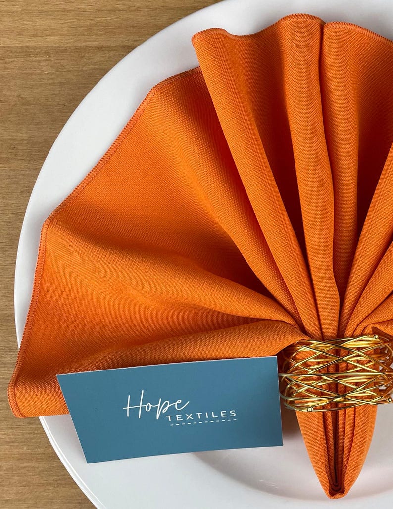 Pode incluir: Guardanapo de linho laranja dobrado em forma de leque e preso com um anel de guardanapo de arame dourado. O guardanapo est&aacute; sobre um prato branco com um cart&atilde;o que diz "Hope Textiles".