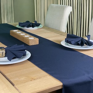 May include: A dark blue table runner with a wooden candle holder in the centre. The runner is placed on a light wood table with white plates and dark blue napkins.