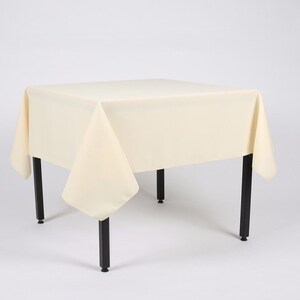 May include: A cream-coloured tablecloth draped over a square table with black legs. The tablecloth has a smooth, even texture and hangs down on all sides of the table.