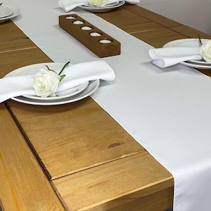 May include: White table runner on a wooden table with white plates, napkins, and flowers. A wooden candle holder with five candle holders is in the centre of the table runner.