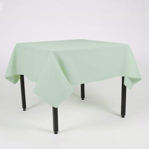 May include: A square, pale green tablecloth on a black metal table. The tablecloth is a solid colour and covers the table, with the corners hanging down. The table has four black legs. The table is approximately 76 cm high.