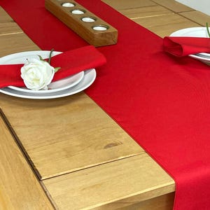 May include: A red table runner on a wooden table with white plates and napkins. A wooden candle holder with five candle holders is on the table runner.